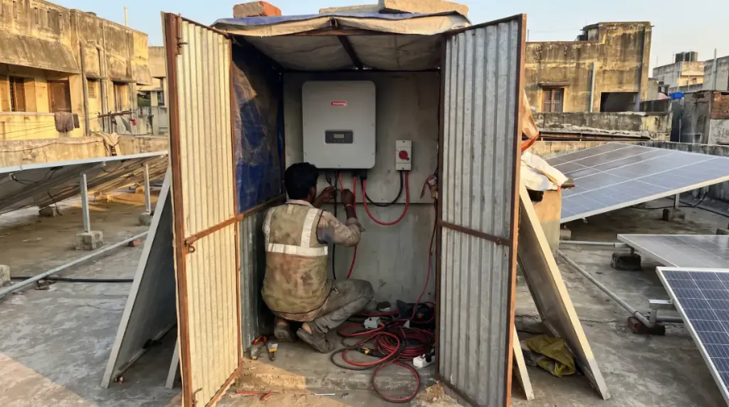 Solar installers at work on a rooftop in Vyasarpadi — an electrician wiring a string inverter to a DC isolator inside a small terrace shed